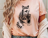 "Raccoon Drinking Iced Coffee" design on a peach unisex t-shirt