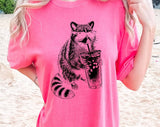 "Raccoon Drinking Iced Coffee" design on a pink watermelon Comfort Colors t-shirt