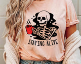 "Staying Alive Coffee Skeleton" design on a peach unisex t-shirt