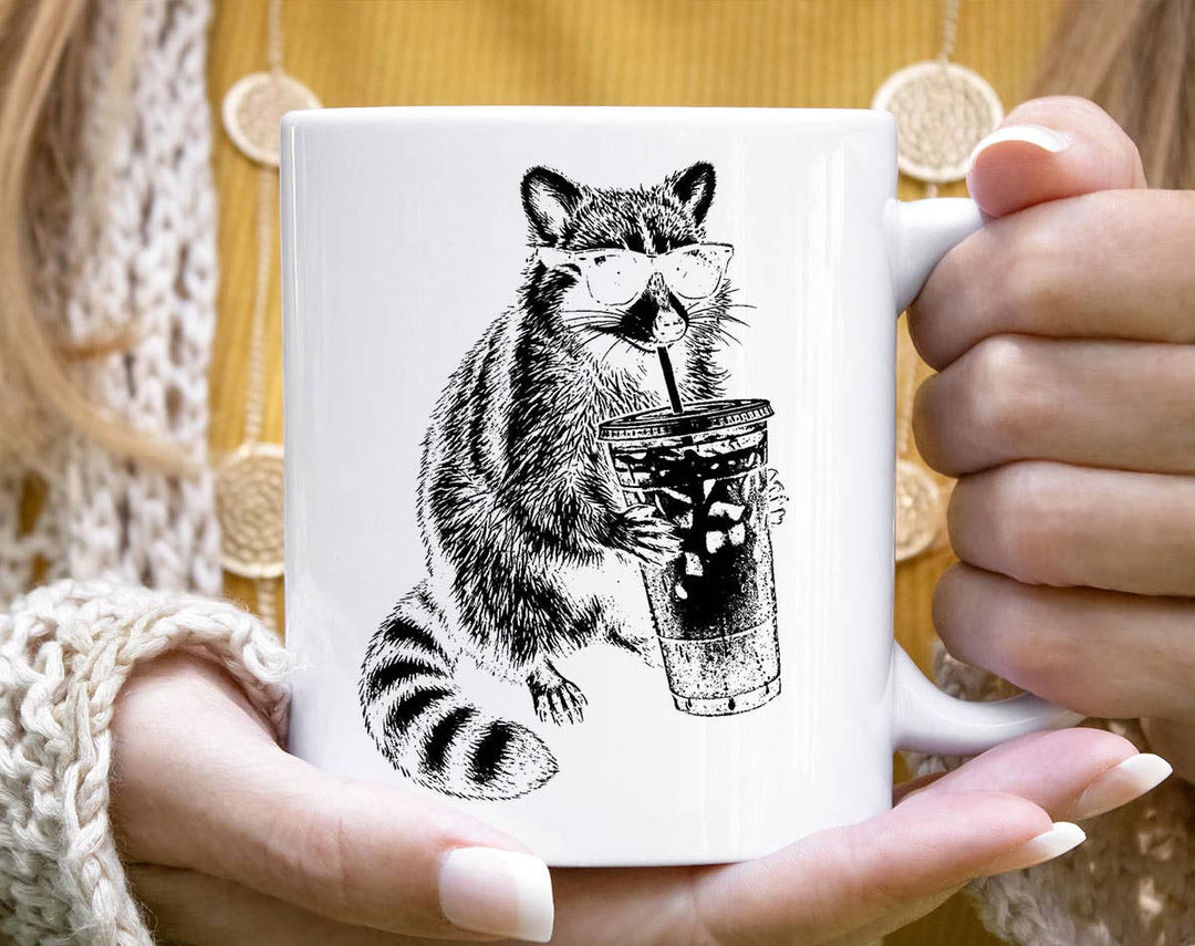 "Raccoon Drinking Iced Coffee" design on a white ceramic coffee mug