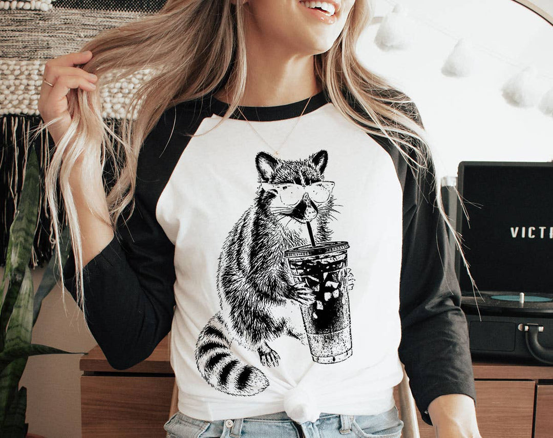 "Raccoon Drinking Iced Coffee" design on a white baseball raglan tee with black sleeves